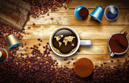 Tazza di caffè vista dall’alto con mappa del mondo sulla superficie, circondata da chicchi, capsule, cialde e polvere: rappresentazione del confronto tra caffè in grani e in capsule.